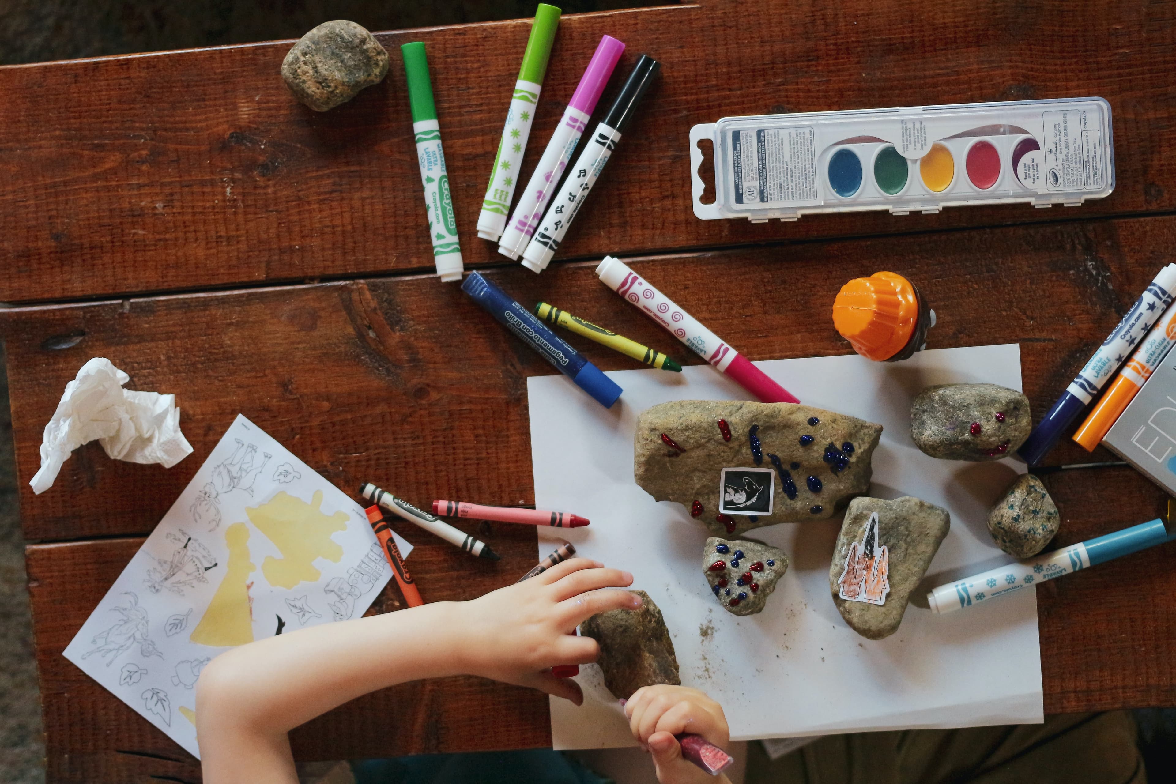 manos de un niño o niña pintando pequeñas piedras con temperas, fibras y crayones sobre una mesa
